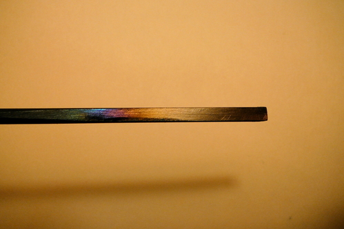 Chisel Tempering
This is a 1/8-inch chisel made from music wire.  To begin, I cut a 6-inch length using a high-speed rotary tool and cut-off disk.  I heated the blank to glowing red and let it cool slowly (annealing).  Then, I was able to shape it with ordinary files.  I shaped it to half thickness for a few inches on one end (half-round cross section), with a bevel tapering from the round side to the end.

After shaping, I heated the working end to glowing red with a torch and plunged it into a can of oil.  I polished the flat surface and slowly heated it over a gas flame until it turned a deep straw-bronze color (tempering).  You can see some purples and blues toward the left (handle) side in the photo.  The steel is softer there, which is fine, because you don't want it to be brittle.

Actually, I messed up the temper the first time, and had to heat treat it again.  Fortunately, you can do that!
Keywords: chisel toolmaking tempering